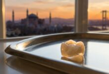 A photorealistic, ultra-detailed macro shot of a perfect E-Max dental crown resting on a sterile surface, with the beautiful, blurred skyline of Istanbul, Turkey visible in the background during a warm sunset.