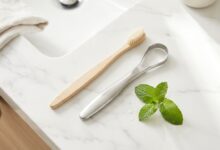 A top-down, photorealistic view of oral hygiene items on a white marble surface, including a bamboo toothbrush, a metal tongue scraper, and a fresh sprig of mint, symbolizing a routine for fresh breath.