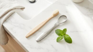 A top-down, photorealistic view of oral hygiene items on a white marble surface, including a bamboo toothbrush, a metal tongue scraper, and a fresh sprig of mint, symbolizing a routine for fresh breath.