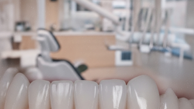 Primer plano de una boca con encías rosadas, sanas y perfectamente contorneadas alrededor de dientes blancos y brillantes, sobre un fondo borroso de una clínica dental moderna y luminosa que sugiere un entorno profesional y de alta calidad.