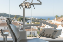 Interior de una clínica dental moderna y luminosa en España, mostrando una silla dental ergonómica y equipos de alta tecnología. A través de una gran ventana, se vislumbra un paisaje urbano o costero español difuminado. La imagen transmite calma y tecnología, sin texto.