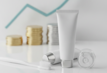Una composición fotorealista de un cepillo de dientes blanco, pasta dental y hilo dental sobre una superficie limpia. En el fondo, elementos financieros borrosos como una pila de monedas simbolizan la prevención dental y el ahorro de costes.